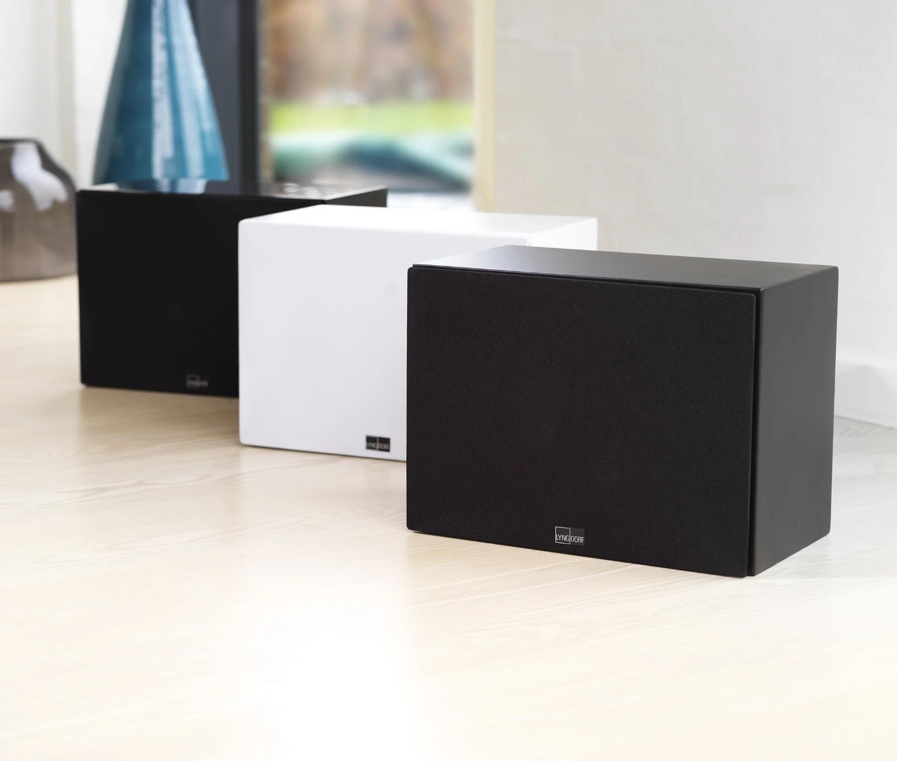 Three Lyngdorf BW-3 subwoofers stand side by side on a beautiful wooden floor, with a window overlooking a garden in the background.