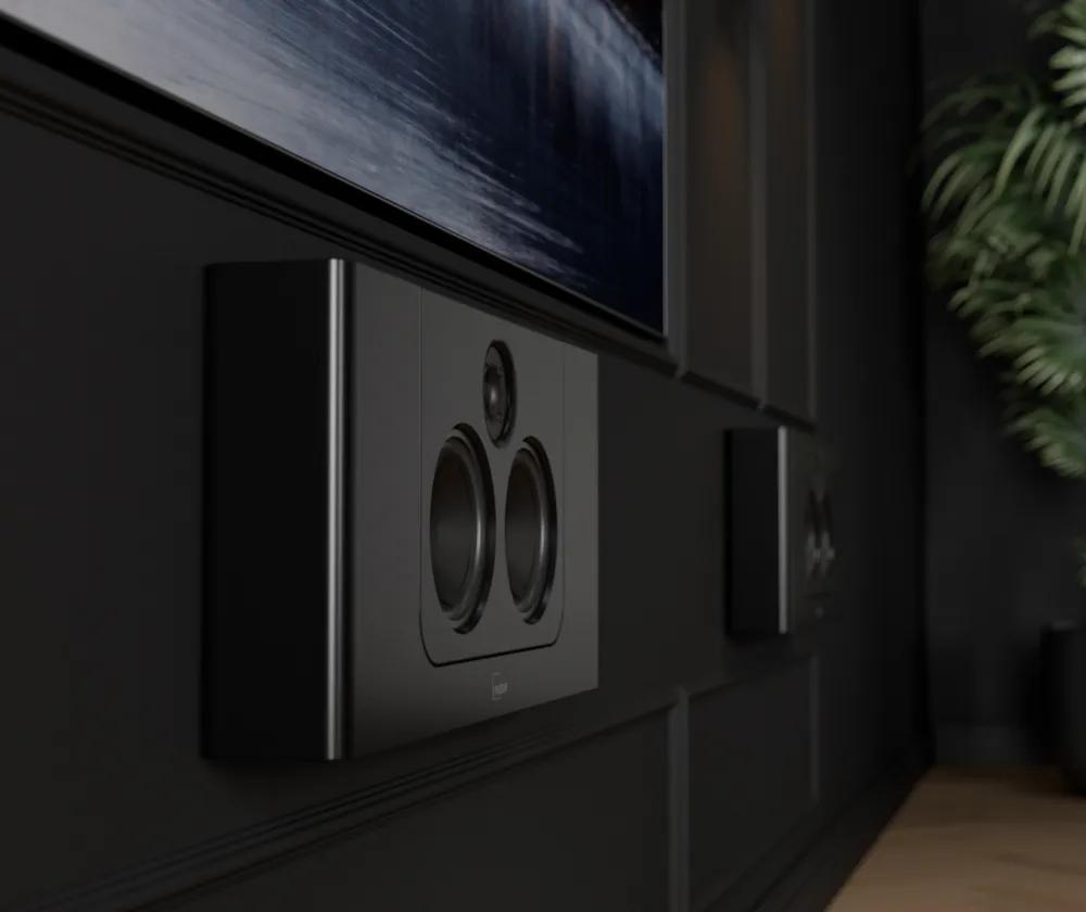 The Lyngdorf LCR-2 speakers are mounted on a black wall; the décor is entirely black, and part of a large television is visible
