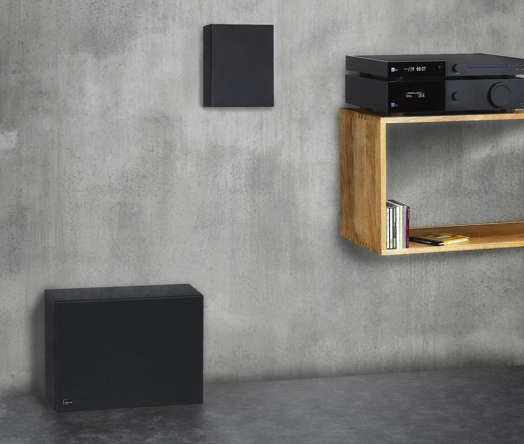 Part of a light grey wall, a black Lyngdorf MH-2 loudspeaker hangs on the wall, a black BW-2 subwoofer stands on the floor, and to the right, on a suspended pedestal, are a CD-2 player and a TDAI-2170 amplifier.