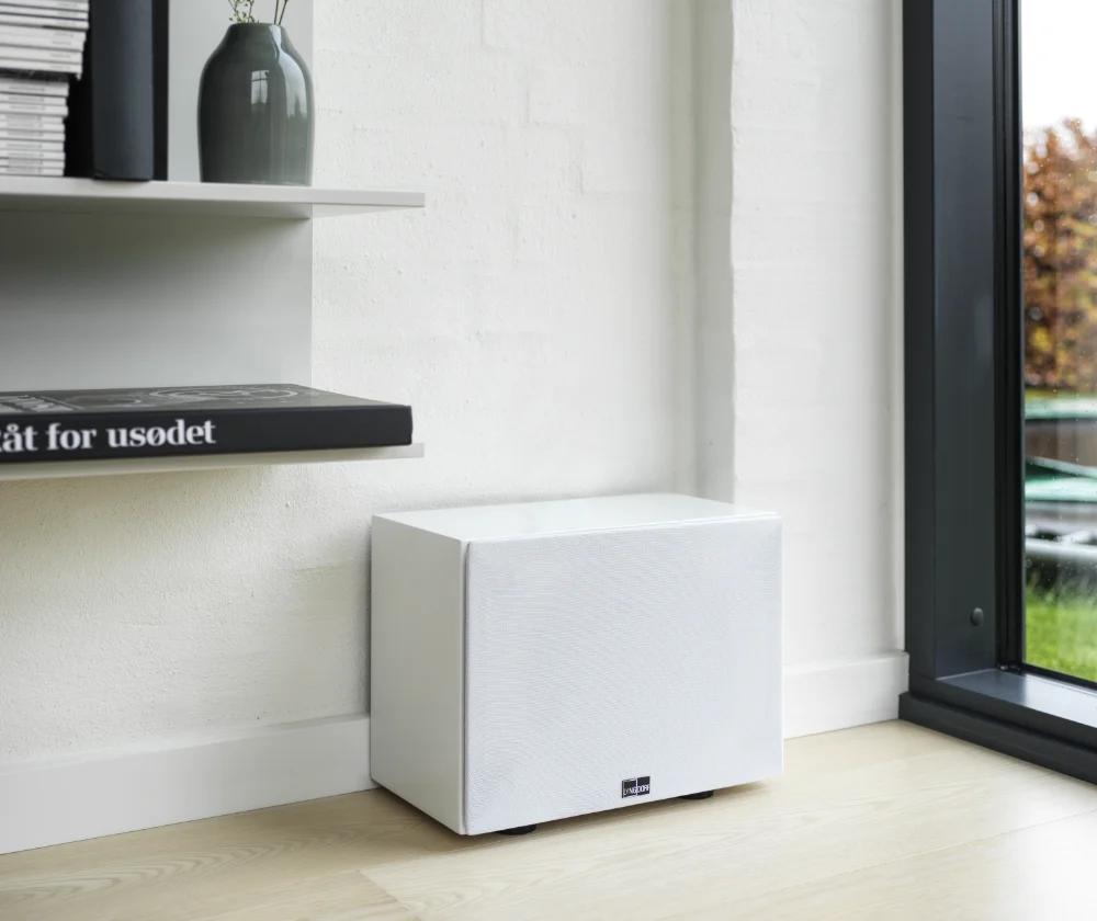 The Lyngdorf BW-3 woofer stands near the wall on a beautiful wooden floor, to the right of a large window/door leading to the garden.