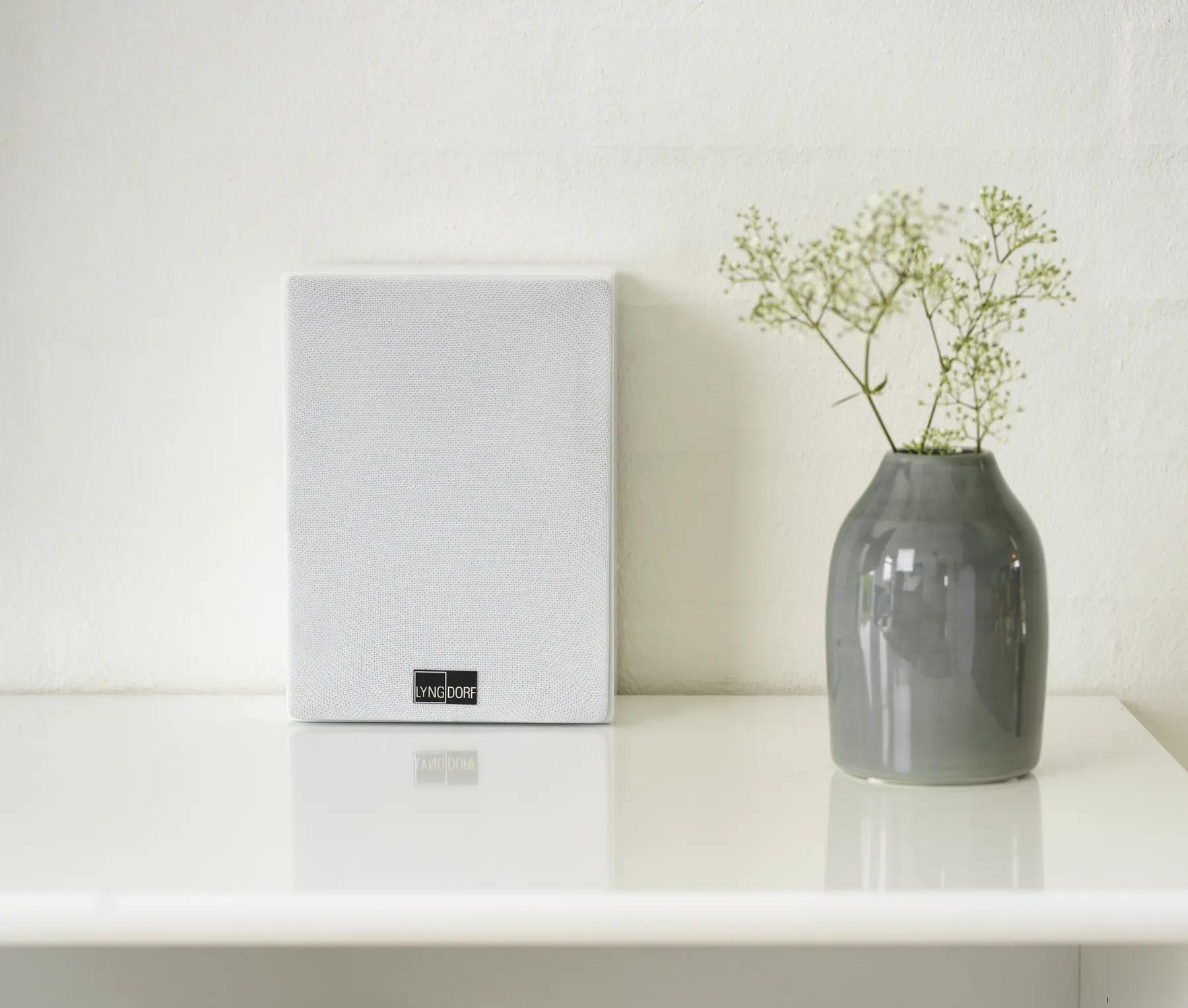 The Lyngdorf MH-3 loudspeaker in white stands on a pedestal, allowing you to appreciate its compact size, which is comparable to the small vase standing next to it and the plate on the shelf below.