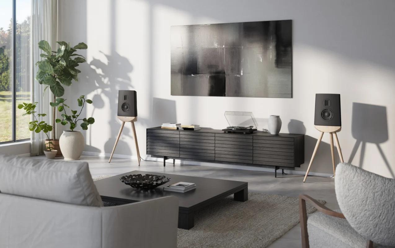 In the room, against the wall, there are two black Cue-100 speakers on wooden stands, with a cabinet containing a vinyl record player between them