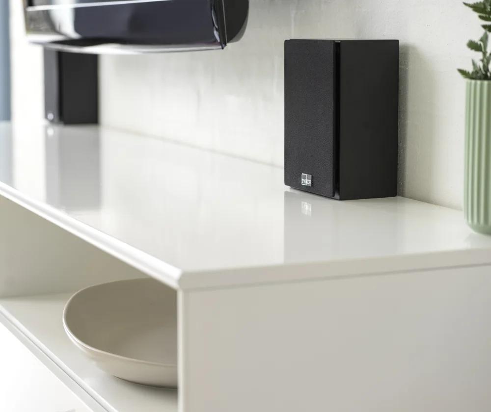 The Lyngdorf MH-3 loudspeakers in black stand on a TV stand, allowing you to appreciate their compact size.