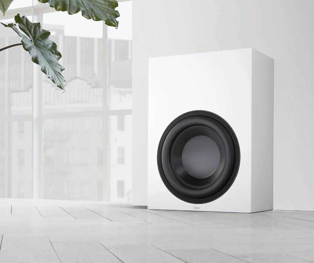 White BW-2 subwoofer against the wall, with the city visible in the floor-to-ceiling window on the left, as well as a beautiful green plant.