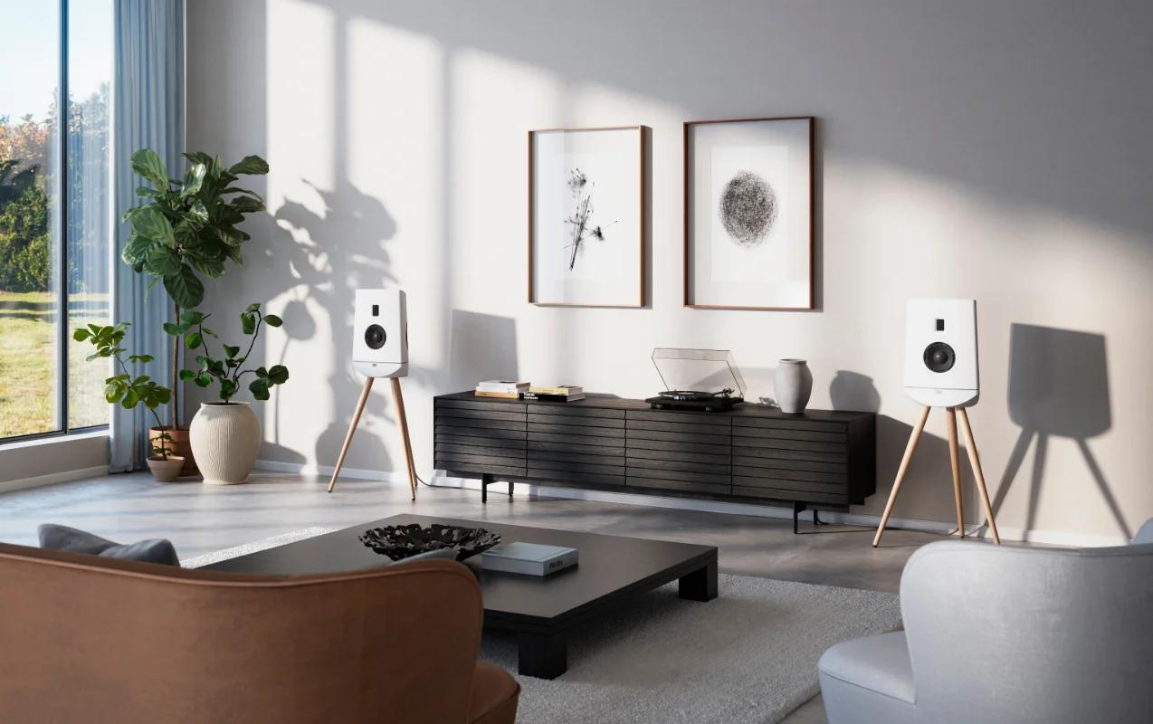 In the room, against the wall, there are a pair of white Lyngdorf Cue-100 speakers on wooden stands, with a cabinet containing a vinyl record player between them