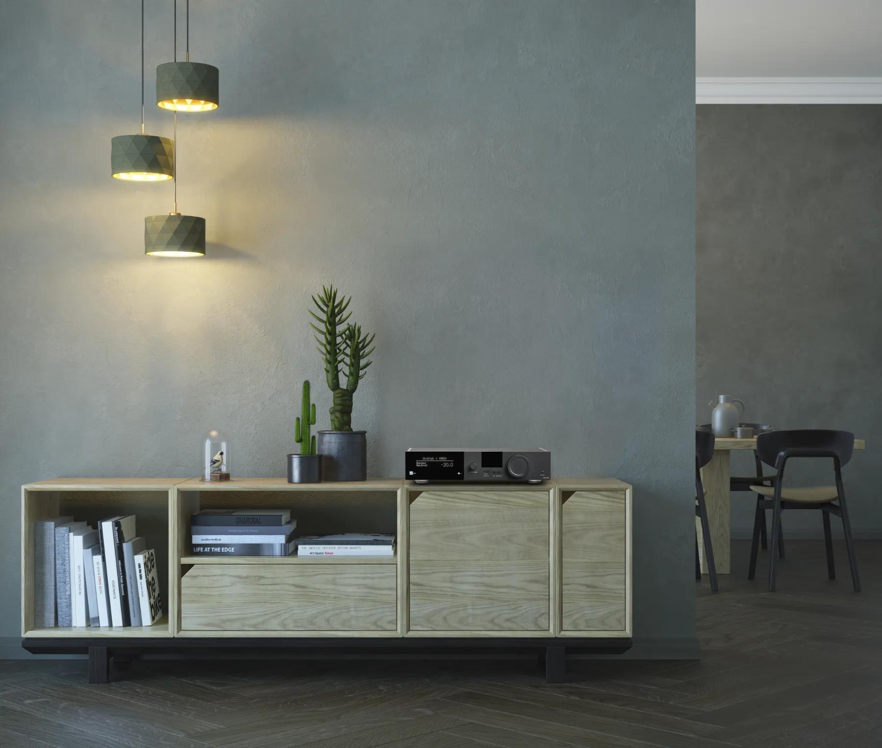 Part of a room with grey walls and dark parquet flooring, a geometric light wood cabinet stands against the wall, with a TDAI-3400 integrated streaming amplifier on the top shelf; beautiful succulents stand nearby, books on the shelves below; in the background, in the adjacent space, a stylish wooden table and chairs can be seen