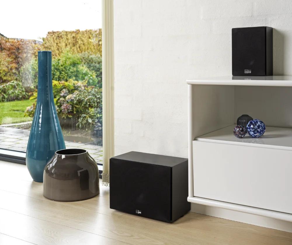 The Lyngdorf MH-3 loudspeaker in black stands on a pedestal, next to which is a black BW-3 subwoofer, with a beautiful garden visible through the large window in the background.