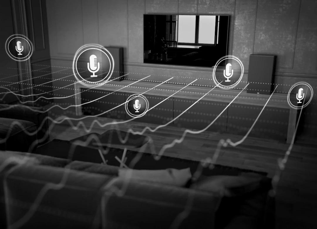 Кімната із диваном та телевізором на стіні, з обох боків телевізора на тумбі встановлені два гучномовці, від яких графічно проведені зображення звукових хвиль в бік дивана. На хвилях зображені значки мікрофонів, які уособлюють вимірювальні мікрофони системи корекції акустики приміщення.